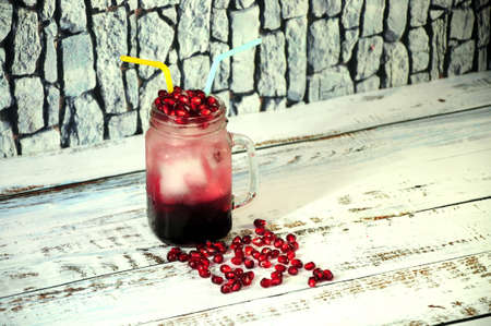 A glass mug of pomegranate juice with ice and a straw is standing on a wooden table against a stone...