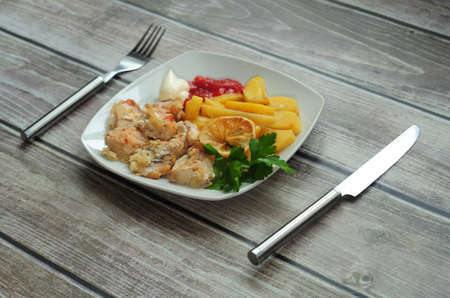 A plate with baked chicken meat and potatoes with sauce, next to a knife and fork. Close-up.