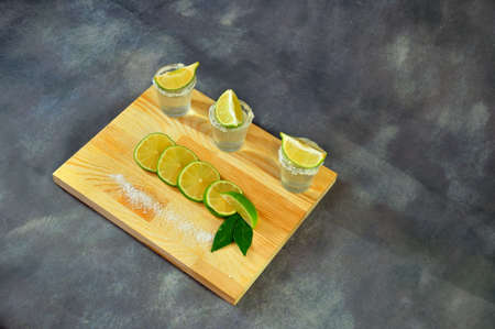 A rectangular wooden stand with three glasses of tequila with lime slices and salt on a gray...