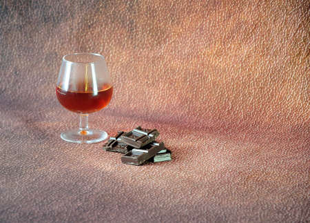 A glass of cognac and a few pieces of chocolate on a brown abstract background. Close-up.