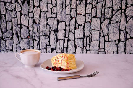 A white ceramic cup of fresh cappuccino with foam, a plate with a slice of puff cake and cherry and...