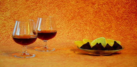Two glasses with cognac and a plate with lemon slices and chocolate candies on a brown abstract...