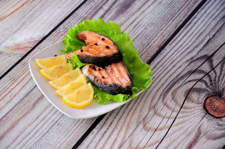 Two grilled pink salmon steaks on a lettuce leaf lie on a plate with lemon slices on a wooden table....