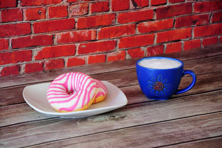 A plate with two donuts in pink strawberry glaze and a blue ceramic cup with cappuccino is on a wooden table against a red brick wall. Close up.の写真素材