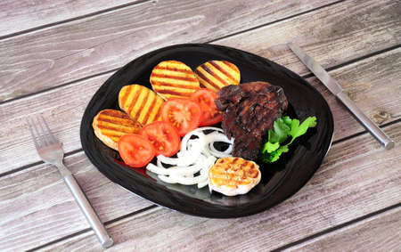 Grilled beef steak, fresh tomatoes and onions, baked head of garlic and potatoes. Close-up.