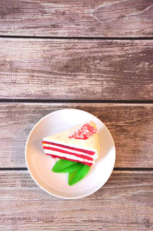 A piece of red velvet cheesecake decorated with mint leaves on a white plate stands on a wooden table. Close-up.の写真素材