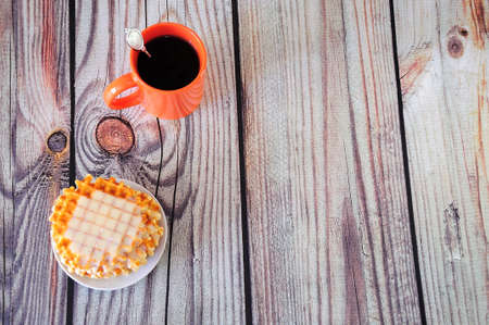 An orange ceramic mug of hot coffee and a plate with Parisian waffles and creamy syrup. Close-up.