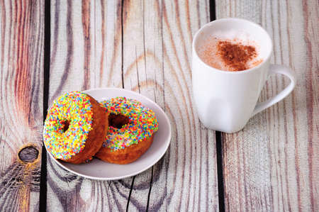 White mug of cappuccino with cinnamon topping and a plate with two donuts in multi-colored topping on a wooden table. Close-up.の写真素材