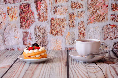 Sponge honey cake with cream, cherry and strawberries stands on a wooden table, next to a mug of tea. Close up.の写真素材