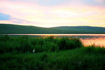 The setting sun has painted pink on the surface of the lake, surrounded by hills and with...