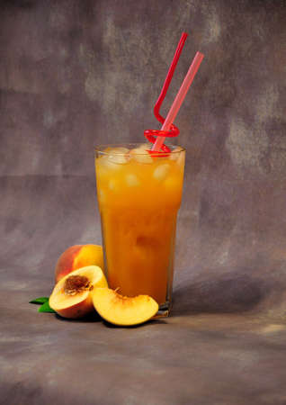 Peach juice in a tall glass with ice and straws, next to pieces of fruit on a gray background. Close-up.の写真素材