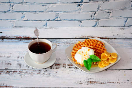 A cup of tea and a plate of waffles, protein cream and banana slices sit on a wooden table against a brick wall. Close-up.の写真素材