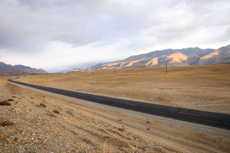 New two-lane asphalt road running along the power line through the autumn steppe surrounded by mountains in the early morning. Chuisky tract, Altai, Siberia, Russia.の写真素材