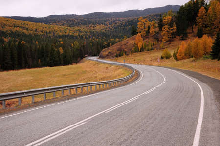 An asphalt two-lane road with fences turns at the foot of the mountain in a picturesque autumn...