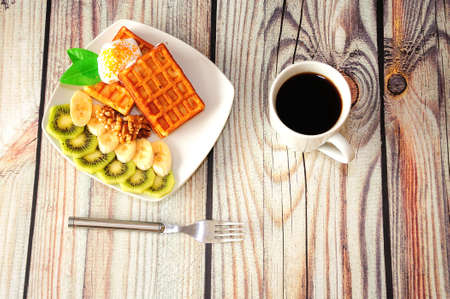 A plate of waffles with ice cream in sprinkles, slices of kiwi and banana, next to a cup of black...