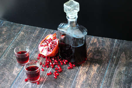 A glass decanter and two glasses of pomegranate liqueur on a wooden table, next to a ripe fruit and a handful of seed. Close-up.の写真素材