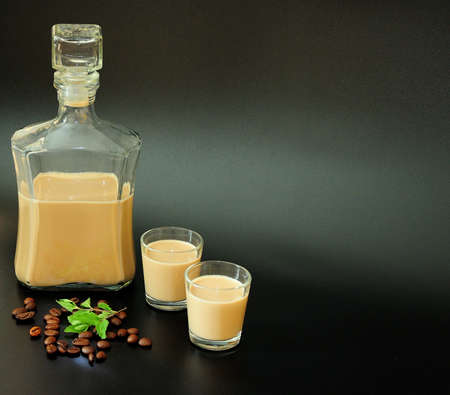 Creamy coffee liqueur in a bottle and two glasses, next to a handful of roasted coffee beans and a sprig of fresh mint on a black background. Close-up.の写真素材