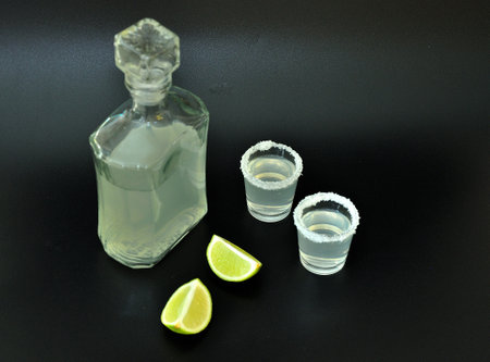 Tequila, a strong homemade alcohol in a glass bottle and two glasses with salt on the edge, next to it are ripe lime pieces on a black background. Close-up.の写真素材