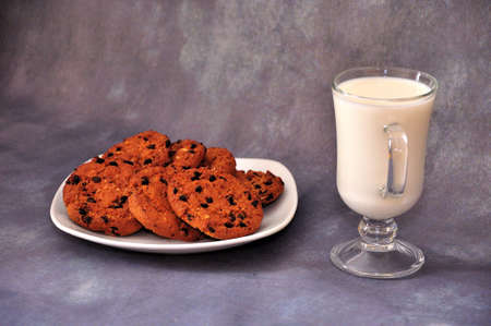 A plate with several oatmeal cookies with chocolate and a glass of milk on a gray isolated...