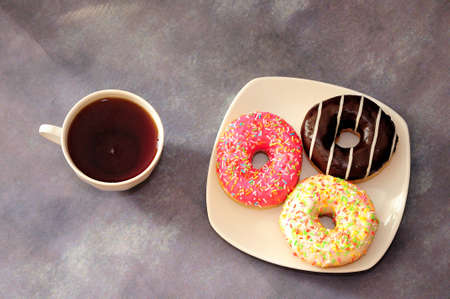 Three donuts in multicolored glaze on a plate and a cup of black tea. Top view, flat lay.の写真素材