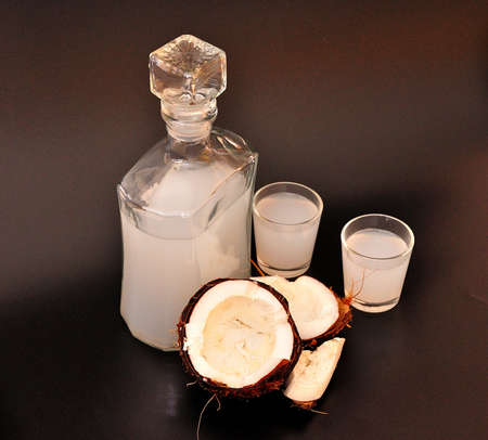 Coconut liqueur in two glasses and a glass decanter, next to a broken nut on a black background. Close-up.の写真素材