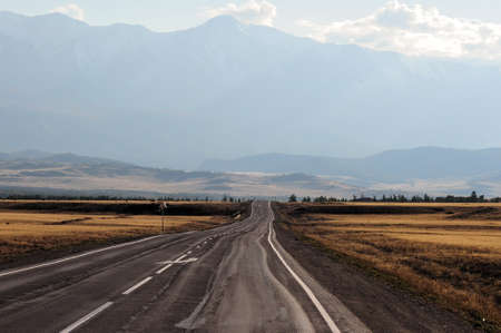 A winding road going through the autumn steppe to meet hills and snow-covered mountain peaks. Chuisky tract, Altai, Siberia, Russia.の写真素材
