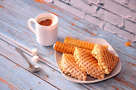 A cup of hot cappuccino, two cubes of sugar and a plate with different waffles on a wooden table....