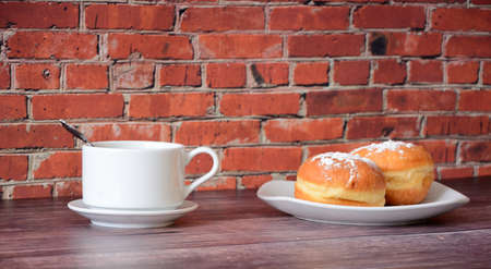 Two fresh donuts sprinkled with powdered sugar and a cup of coffee stand on a wooden table against the wall. Close-up.の写真素材