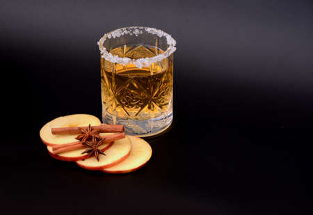 Crystal glass of apple cider with sugar on the rim, pieces of ripe fruit, cinnamon sticks and anise on a black background. close-up.の写真素材