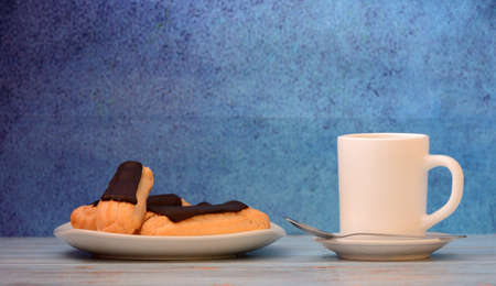 A plate with several eclairs in chocolate glaze and a cup of fresh coffee on a wooden board. front view.の写真素材