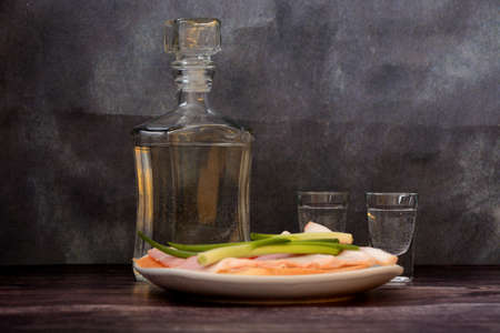A bottle of vodka, two glasses and a plate with two sandwiches with bacon and green onions on a wooden table. close-up.の写真素材