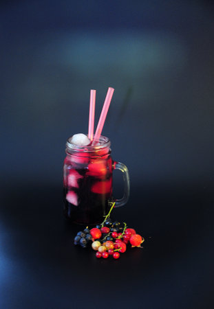 Berry juice in a glass mug with ice and tubes on a black background, next to a guest of various garden berries. close-up.の写真素材