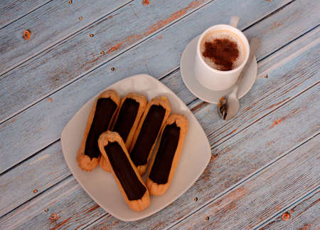 A plate with several eclairs in chocolate glaze and a cup of black coffee on a wooden table. Top...