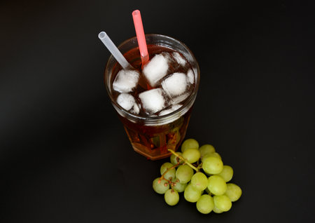 A tall glass of grape juice with ice and straws on a black background, next to a bunch of ripe green fruits. Top view, flat lay.の写真素材