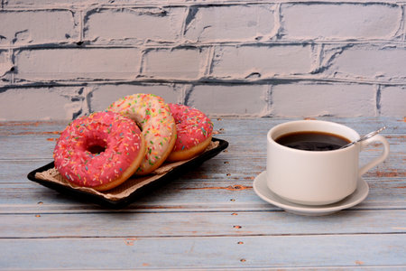 A plate with three glazed donuts with sprinkles and a cup of hot black coffee on a wooden table. close-up.の写真素材