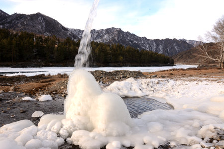 A small stream flows onto an ice block in a picturesque winter valley surrounded by snow-capped mountains on the banks of a beautiful river. Katun river, Altai, Siberia, Russia.の写真素材