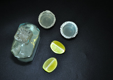 Ripe lime slices, a bottle of tequila and two glasses with salt around the edges on a black background. Top view, flat lay.の写真素材