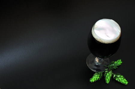 A glass of dark beer with thick white foam and ripe hops on a black background. close-up.