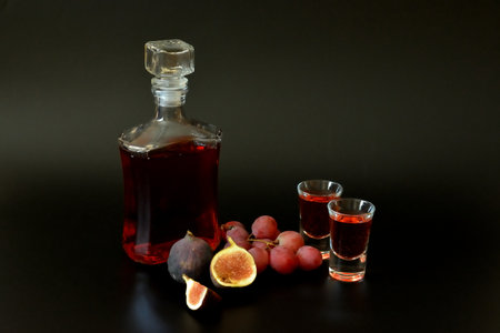 An antique glass decanter and two glasses of homemade liqueur on a black background, next to pieces of ripe figs and a bunch of red grapes. close-up.の写真素材