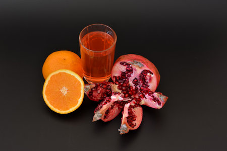 A transparent glass cup of a mixture of freshly squeezed tropical fruits on a black background, next to it is a half of a ripe orange and a broken pomegranate fruit. close-up.の写真素材