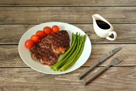 Grilled beef steak, cherry tomatoes and boiled asparagus on a wooden table, next to a gravy boat and cutlery. close-up.の写真素材