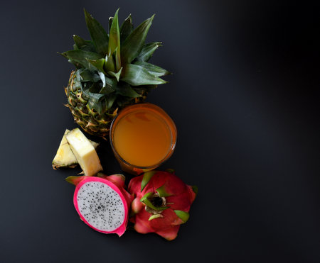 A glass of fruit juice on a black background, next to pieces of pineapple and a cut ripe pitaya fruit. Top view, flat lay.の写真素材