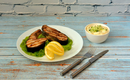 A plate with two salmon steaks on a lettuce leaf with lemon, sauce and cutlery on a wooden table....