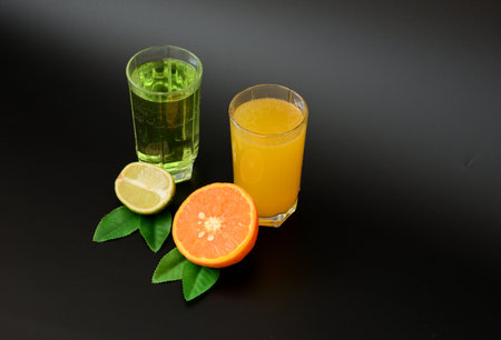 Two glasses with different citrus juices on a black background, next to half a ripe lime and an orange with leaves. close-up.の写真素材
