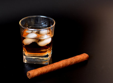 A glass of whiskey with ice cubes and a large Cuban cigar on a black background. Close-up.の写真素材