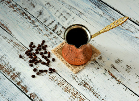 Copper Turk with a hot drink on a jute napkin and a scattering of roasted coffee beans on a light wooden table. Close-up.の写真素材