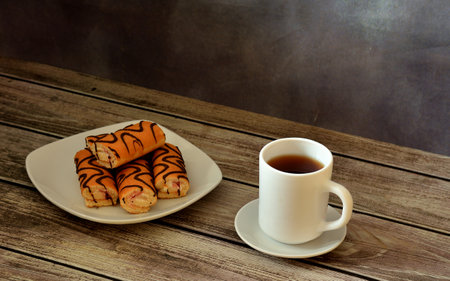A plate with several mini sponge rolls in chocolate glaze and a cup of hot tea on a wooden table....