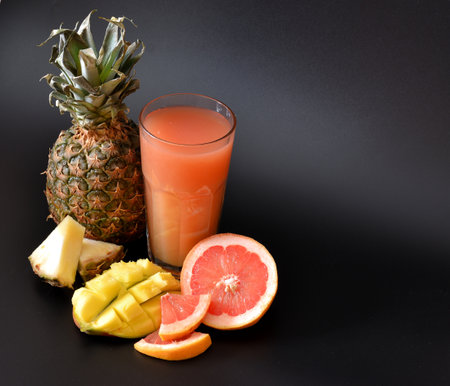 A tall faceted glass of a mixture of tropical fruits on a black background, next to a ripe pineapple, grapefruit and mango. Close-up.の写真素材