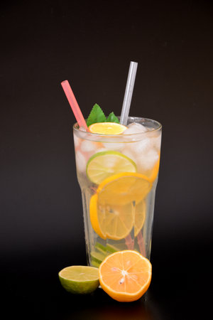A tall faceted glass of refreshing lemonade with ice on a black background, next to pieces of ripe orange and lime. Vertical arrangement.の写真素材