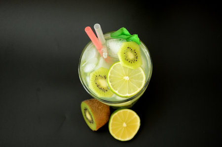 A tall glass of refreshing fruit juice with ice on a black background, next to slices of lime and a ripe kiwi fruit. Top view, flat lay.の写真素材